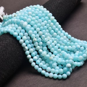 Pode incluir: Um fio de contas de água-marinha azul claro. As contas são redondas e lisas, e são enfiadas em um cordão branco. As contas têm aproximadamente 6 mm de diâmetro.