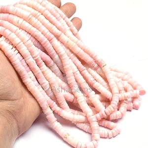 Puede incluir: Una colección de cuentas rosas planas en forma de disco. Las cuentas están ensartadas, creando múltiples hebras. El color es un rosa pastel suave. Las cuentas se sostienen en una mano sobre un fondo blanco. Estas cuentas son ideales para la fabricación de joyas.