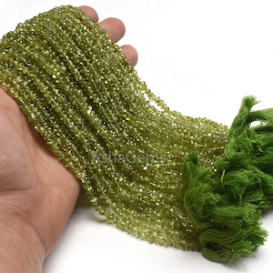Pode incluir: Um fio de contas de peridoto verde, facetadas e enfiadas em um fio. As contas são pequenas e de tamanho uniforme. O fio é longo e está sendo segurado em uma mão.
