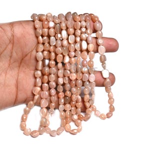 May include: Strands of peach-colored, polished gemstone beads. The beads are oval-shaped and strung together, with variations in color and translucency. The beads are held in a hand against a white background.