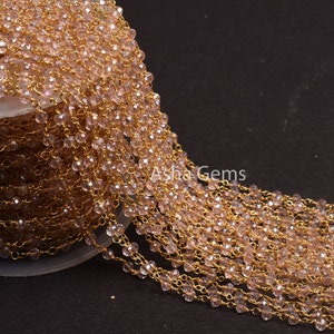 Puede incluir: Primer plano de una cadena dorada con cuentas de cristal rosa. La cadena está enrollada y drapeada sobre una superficie. Las cuentas son facetadas y tienen un brillo sutil.