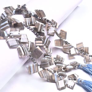 May include: A necklace with square, gray and white striped beads. The beads are strung together with small silver beads. Light blue tassels hang from the ends of the necklace.