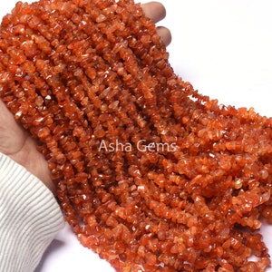 May include: A strand of orange carnelian chip beads. The beads are small and irregular in shape, and they are strung on a clear thread. The beads are arranged in a loose, flowing pattern.