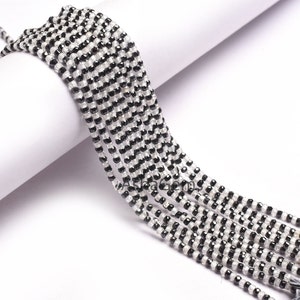 May include: A collection of multiple strands of small, faceted, round beads arranged in alternating rows of clear, translucent white and opaque black. The beads are strung together in parallel lines, creating a high-contrast, monochromatic pattern.