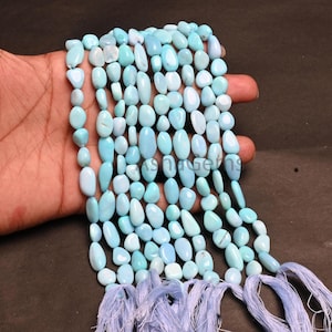 May include: Strands of light blue gemstone beads, each with a unique oval shape. The beads are strung together, with a tassel of matching blue threads at the bottom. The beads are held in a hand.