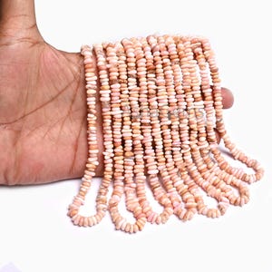 Puede incluir: Múltiples hilos de pequeñas cuentas de piedra de color rosa pálido y melocotón, de forma irregular, dispuestas en líneas paralelas. Las cuentas tienen una forma lisa, redondeada y aplanada, creando una textura uniforme en los hilos.