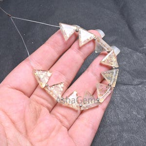 Puede incluir: Un hilo de cuentas triangulares transparentes con inclusiones de purpurina dorada. Las cuentas están ensartadas en un fino alambre plateado. Las cuentas se sostienen en una mano sobre un fondo oscuro. El texto "AshaGem" es visible.