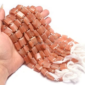 Puede incluir: Hilo de cuentas rectangulares de color melocotón con espaciadores plateados. Las cuentas se sostienen en una mano, con borlas blancas visibles al final. Las cuentas tienen un aspecto pulido y ligeramente translúcido.