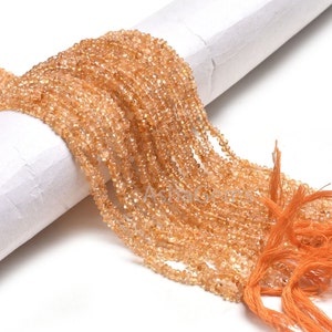 以下が含まれることがあります： 小さく、カットされた、明るいオレンジ色のビーズの連なり。ビーズは紐でつながれ、白い円筒形の物体にかけられています。ストランドの端はオレンジ色の糸で結ばれています。ビーズはキラキラと光る半透明の外観をしています。