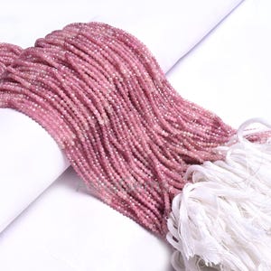 Puede incluir: Una hebra de cuentas de piedras preciosas rosas, de aproximadamente 2 mm de diámetro, enhebradas en un hilo blanco. Las cuentas están dispuestas en una pila suelta, con un borla blanca en la parte inferior.