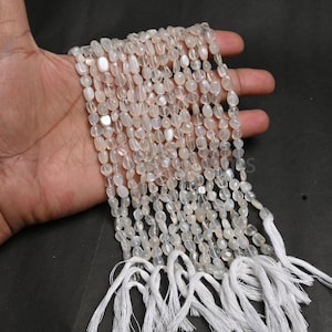 May include: Strands of off-white, oval-shaped beads with a pearlescent sheen. The beads are strung together and tied with white tassels. The beads are held in a hand against a dark background. These beads are suitable for jewelry making.