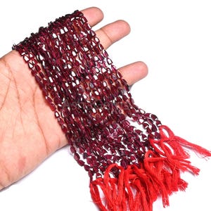 Puede incluir: Hebras de cuentas de piedras preciosas facetadas de color rojo intenso. Las cuentas tienen forma de lágrima y están ensartadas, con una borla roja brillante en el extremo. Las cuentas se sostienen en una mano sobre un fondo blanco.