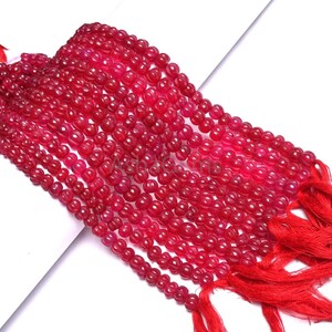 May include: Strands of small, round, deep red beads, possibly gemstones, are displayed against a white background. The beads are strung together, with a tassel of red threads at the end of each strand. The beads have a glossy finish.