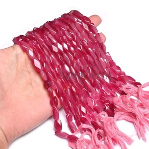 Puede incluir: Un hilo de cuentas facetadas, translúcidas y de color magenta con un acabado pulido. Las cuentas son alargadas y de forma irregular, ensartadas en un hilo de color rosa claro que termina en borlas sueltas y deshilachadas en el extremo.