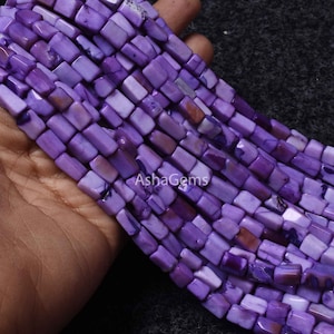 Puede incluir: Primer plano de hebras de cuentas de piedras preciosas moradas. Las cuentas son rectangulares con bordes redondeados y un acabado pulido. El color varía de morado claro a oscuro con algunas inclusiones marrones. Las cuentas están ensartadas, listas para hacer joyas.