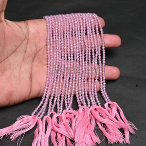 Puede incluir: Hebras de cuentas de piedras preciosas de color rosa claro con corte facetado. Las cuentas se mantienen unidas y se terminan con borlas rosas. Las cuentas se muestran sobre un fondo oscuro.