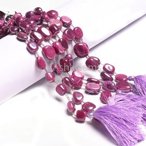 May include: A strand of pink ruby gemstone beads with a silver spacer between each bead. The beads are oval shaped and have a smooth, polished finish. The strand is finished with two purple tassels.