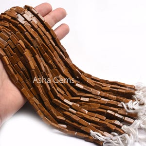 Puede incluir: Una hebra de cuentas de piedras preciosas marrones, cada cuenta tiene forma rectangular. Las cuentas están enhebradas en un cordón blanco y se muestran en una mano.