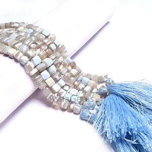 Könnte beinhalten: Eine Strähne aus hellblauen und weißen quadratischen Edelsteinperlen mit blauen Quasten an den Enden.