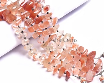 May include: Strands of raw, faceted gemstones in shades of orange and peach. The beads are strung together with small black beads and silver-toned findings. Tassels hang from the bottom of the strands. The text "Asha Gems" is visible.