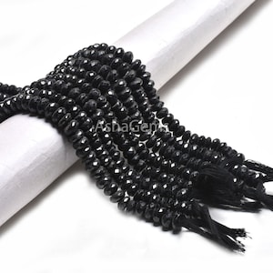 May include: Strands of faceted black beads with a tassel at the end. The beads are draped over a white cylindrical object. The beads are a deep black color and appear to be made of a shiny material. The image has the text "AshaGems".
