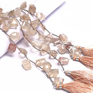 May include: A strand of light brown faceted gemstone beads with silver wire strung between each bead. The beads are in the shape of a rectangle with a pointed top. The strand is displayed on a white surface with two brown tassels.