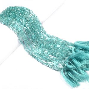 Könnte beinhalten: Eine Strähne aus hellblauen Aquamarin-Edelsteinkugeln, jede Kugel hat eine rechteckige Prismaform. Die Kugeln sind auf einer dünnen Schnur aufgefädelt und haben einen Quasten am Ende.