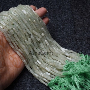 Puede incluir: Una hebra de cuentas de piedras preciosas rectangulares de color verde claro. Las cuentas están enhebradas en un cordón fino y tienen un borla en el extremo. Las cuentas miden aproximadamente 1,2 cm de largo y 0,6 cm de ancho. Las cuentas son translúcidas y tienen un ligero brillo.