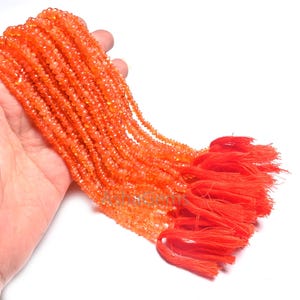 Puede incluir: Una colección de cuentas de piedras preciosas de color naranja, sostenidas en una mano, con una borla naranja brillante en el extremo. Las cuentas son pequeñas y de tamaño uniforme, creando un efecto brillante. La borla está hecha de hilos gruesos y vibrantes de color naranja.