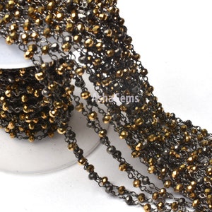 May include: A close-up of a gold and black chain made of faceted beads. The chain is coiled on a white spool.