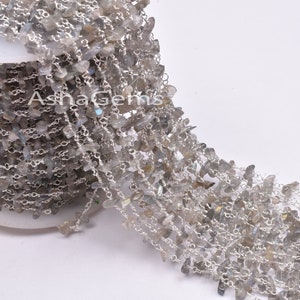 Könnte beinhalten: Eine silberne Kette mit kleinen, unregelmäßigen, grauen Labradorit-Perlen. Die Kette ist aufgerollt und auf einer weißen Oberfläche drapiert.