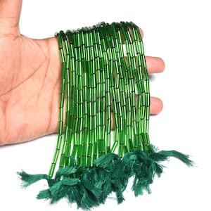 May include: Strands of translucent green glass beads, each cylindrical in shape, are held in a hand. The beads are strung together, with a tassel of green thread at the end of each strand. The beads have a glossy finish and are displayed against a white background.