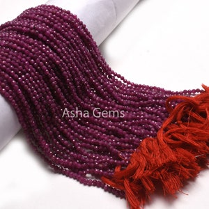 Op de afbeelding: Een streng dieprode robijn kralen, ongeveer 2 mm groot, met een facetgeslepen afwerking. De kralen zijn geregen op een dun draad en hebben een kwast aan het einde. De kralen worden gepresenteerd op een witte achtergrond.