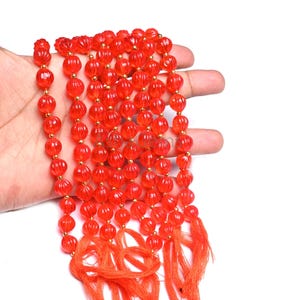May include: A collection of bright orange beads with a ribbed design and gold-coloured spacers. The beads are strung together, forming multiple rows, and are finished with orange tassels. The beads are held in a hand against a white background.