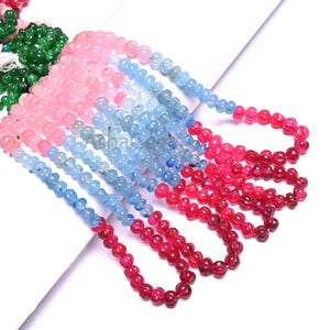 Può includere: Fili di perline colorate di pietre preziose nei toni del rosa, blu e rosso. Le perline rotonde sono infilate per creare collane o bracciali. Sono visibili anche alcuni fili di perline verdi.
