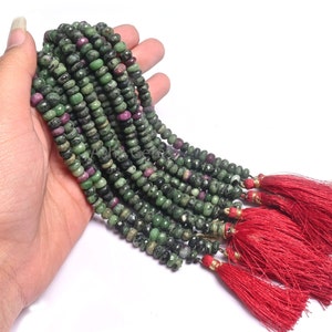 Puede incluir: Una hebra de cuentas de piedras preciosas verdes y rosas con borlas rojas. Las cuentas son redondas y tienen un aspecto moteado.