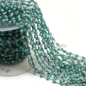 May include: A spool of turquoise blue faceted glass beads strung on a black chain. The chain is made of small, linked metal rings. The beads are uniform in size and shape.