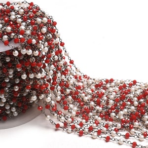 Puede incluir: Una cadena de cuentas con cuentas rojas y blancas alternas. Las cuentas están ensartadas en una cadena de metal oscuro. La cadena está enrollada, lo que sugiere que se vende por metros. Las cuentas rojas son facetadas.