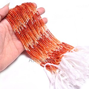 Könnte beinhalten: Eine Strähne aus orangefarbenen, roten und weißen Edelsteinperlen. Die Perlen sind klein und rund und auf einem weißen Faden aufgefädelt. Die Strähne ist mit einem weißen Quast gebunden.