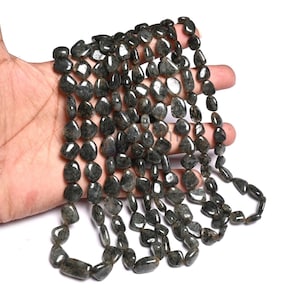 May include: Strands of dark gray tumbled gemstone beads. The beads are irregular in shape and size, with a polished, reflective surface. The beads are strung together, forming multiple strands. The background is white.