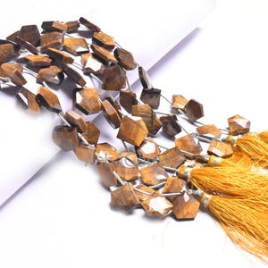 Puede incluir: Una hebra de cuentas de piedra de ojo de tigre marrón y dorado con forma cuadrada facetada. Las cuentas están enhebradas en un alambre plateado y tienen un borla en el extremo.