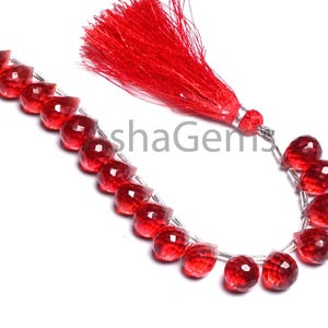 Op de afbeelding: Een streng gefacetteerde, druppelvormige rode edelstenen. De kralen zijn geregen op een zilverkleurige draad, met een rode kwast aan het uiteinde. De edelstenen hebben een glanzende afwerking en zijn in een gebogen lijn gerangschikt.