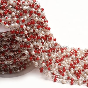 Puede incluir: Primer plano de una cadena de rosario blanca y roja con cuentas plateadas y una cadena plateada. La cadena está enrollada sobre una superficie blanca.