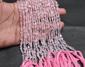 May include: Strands of oval-shaped, translucent rose quartz beads. The beads are a soft pink color and are strung together. Pink tassels are visible at the bottom of the strands. The beads are held in a hand against a dark background.