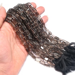 Peut inclure: Un fil de perles rectangulaires en quartz fumé. Les perles sont de couleur marron translucide et sont enfilées ensemble. Les perles sont tenues dans une main sur un fond blanc. Les perles sont prêtes à être utilisées pour la fabrication de bijoux.