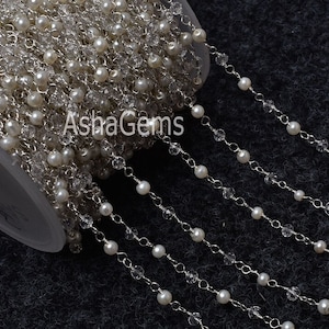 Peut inclure: Une chaîne en argent avec des perles blanches et des perles de cristal transparentes. La chaîne est enroulée sur une surface blanche. Le texte "AshaGems" est visible sur la chaîne.