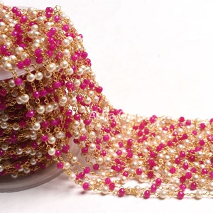 May include: A gold chain with pink and white beads. The chain is made of small, round beads that are strung together in a repeating pattern. The chain is coiled up on a white surface.