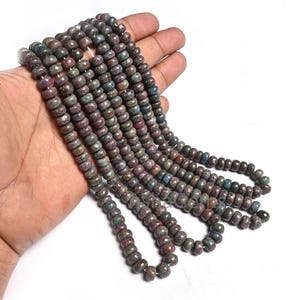 Könnte beinhalten: Stränge aus Edelsteinperlen in Braun-, Grün- und Lilatönen. Die zylindrischen Perlen sind aufgefädelt und bilden eine Halskette oder ein Armband. Die Perlen werden in einer Hand vor weißem Hintergrund gehalten.