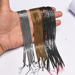 May include: Three bundles of beaded strands in silver, gold, and black are held in a hand. Each strand is composed of small, rectangular beads. The strands are displayed against a white background, showcasing the different colors and textures of the beads.