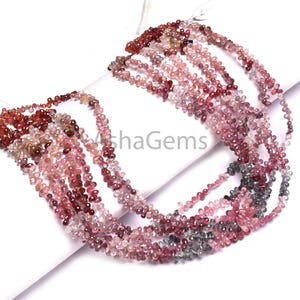 May include: Strands of gemstone beads in shades of pink, red, and gray. The beads are small, teardrop-shaped, and strung together. The image is taken on a white surface. The text "AshaGems" is visible in the image.